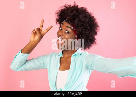 Buschige afroamerikanische junge Frau, die Selfie posiert, gestikulierend V-Sign über rosa Studiohintergrund, lächelnd vor der Kamera. Erfolgreiche Millennial-Frau Stockfoto
