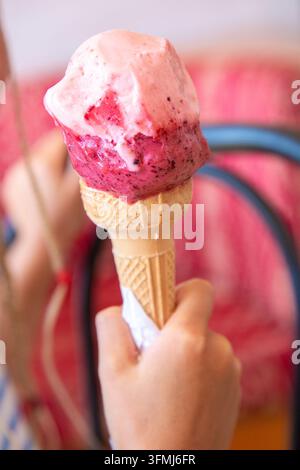Nahaufnahme einer Hand, die ein Eis mit Erdbeere und Beeren hält, mit schmelzenden rosa Spiralen, cremiger Textur, Waffelkegel, weicher verschwommener Hintergrund, verspielt Stockfoto