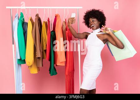 Fröhliche Afrikanische Shopaholic-Frau, Die Selfie Am Telefon Während Des Einkaufs Macht Posiert Mit Shopper Bag In Der Nähe Von Kleiderständer Mit Trendigen Neuen Kleidung Auf Rosa Stud Stockfoto