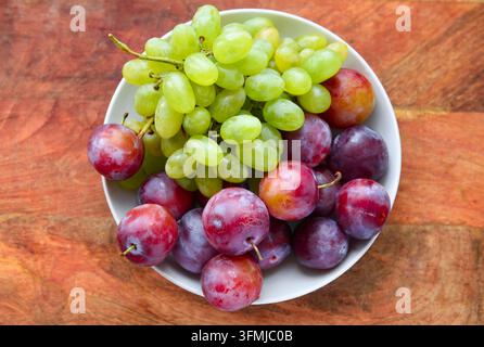 grüne Trauben und Pflaumen auf einem Teller auf einem Holzbrett, Konzept von frischen Früchten und gesunder Ernährung Stockfoto