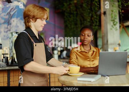 Barista serviert Kaffee an eine schwarze Frau, die in einem Café an einem Laptop arbeitet Stockfoto