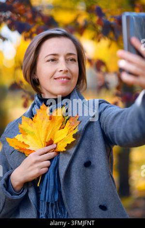 schöne Frau mit herbstgelben Blättern macht ein Selfie auf ihrem Handy, posiert in einem Stadtpark, heller sonniger Tag in der Herbstsaison Stockfoto
