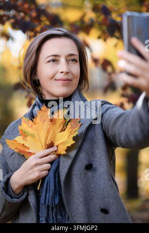 schöne Frau mit herbstgelben Blättern macht ein Selfie auf ihrem Handy, posiert in einem Stadtpark, heller sonniger Tag in der Herbstsaison Stockfoto