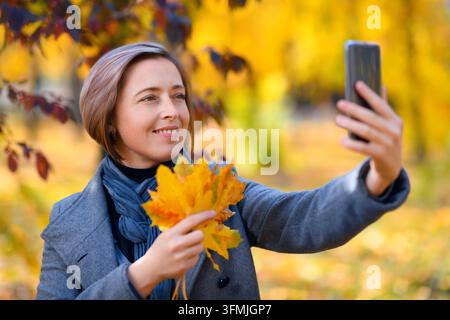 schöne Frau mit herbstgelben Blättern macht ein Selfie auf ihrem Handy, posiert in einem Stadtpark, heller sonniger Tag in der Herbstsaison Stockfoto