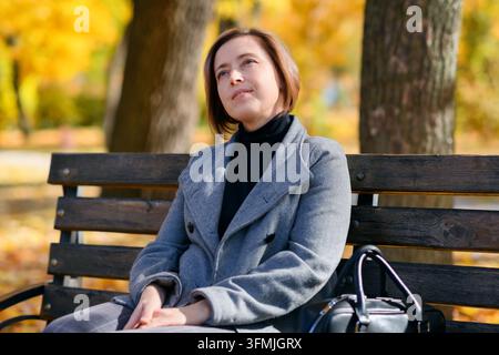 schönes Frauenporträt, sie sitzt auf einer Bank im Herbstpark, Bäume mit gelben Blättern, heller Sonnentag Stockfoto