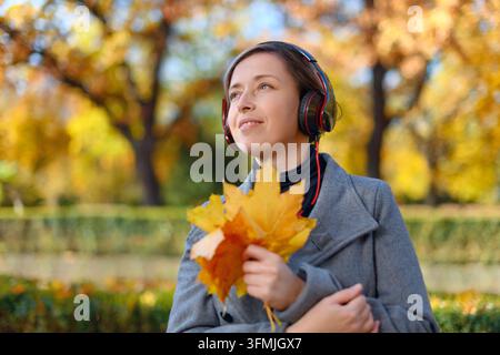 schönes Frauenporträt, sie ist in einem Herbstpark und hört Musik mit Kopfhörern, Bäume mit gelben Blättern, heller Sonnentag Stockfoto