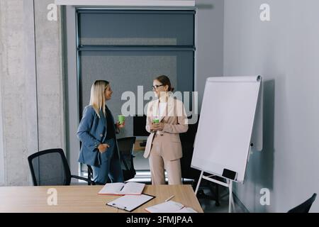 Zwei professionelle Geschäftsfrauen führen ein freundliches Gespräch und trinken Kaffee während einer Pause in einem modernen Bürotreffenraum Stockfoto