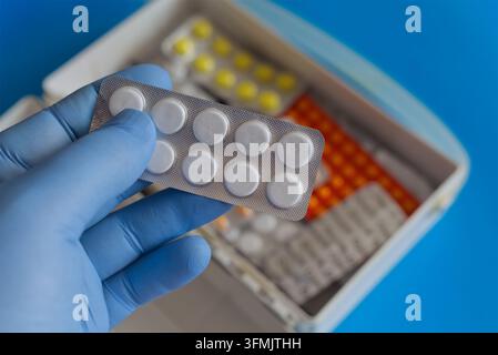 Hand in einem blauen medizinischen Handschuh, die eine Blisterpackung mit weißen medizinischen Pillen hält, vor dem Hintergrund eines offenen Erste-Hilfe-Koffers mit verschiedenen bunten Medikamenten. Stockfoto