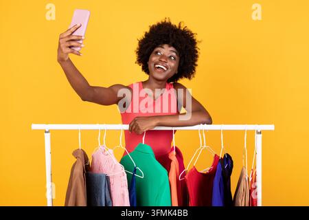 Fröhliche Schwarze Dame, Die Selfie Auf Smartphone Macht, Posiert In Der Nähe Von Kleiderstange Mit Trendigen Neuen Kleidern Über Gelbem Hintergrund Im Studio. Shopping Applicati Stockfoto