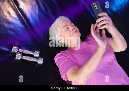 Aktive Seniorin 75+ nutzt Smartphone, während sie sich nach dem Training ausruht. Technologie, Seniorfitness, gesundes Altern und digitaler Lebensstil Stockfoto