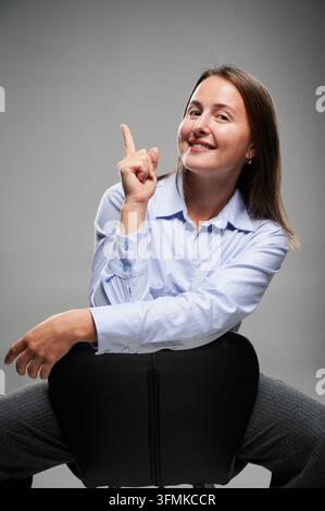 Geschäftsfrau sitzt rückwärts auf dem Stuhl und zeigt nach oben im Studio-Porträt. Junge erwachsene Frau mit braunem Haar, hellblauem Hemd, grauen Hosen, fröhlicher selbstbewusster Ausdruck, professioneller Stil Stockfoto