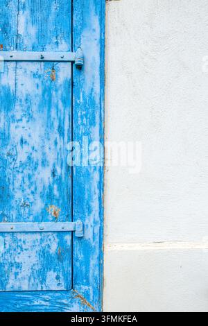 Blauer abblätternder Holzläden, detaillierte rustikale Metallscharniere, strukturierte weiße Stuckwand, minimalistische architektonische Außenansicht Stockfoto