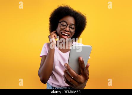 Zufriedene afroamerikanische Dame, die ein Smartphone benutzt und einen Videoanruf tätigt, in die Handykamera lächelt, vor gelbem Hintergrund posiert. Aufgeregte schwarze Frau taki Stockfoto