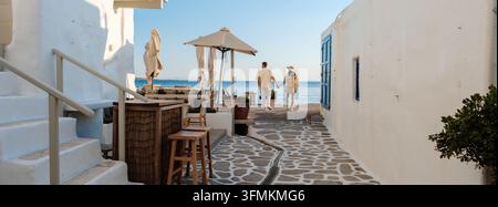 Ein Paar geht Hand in Hand entlang der Küste von Naousa in Paros, Griechenland, umgeben von bezaubernden weiß getünchten Gebäuden und dem goldenen Licht des Sonnenuntergangs. Stockfoto