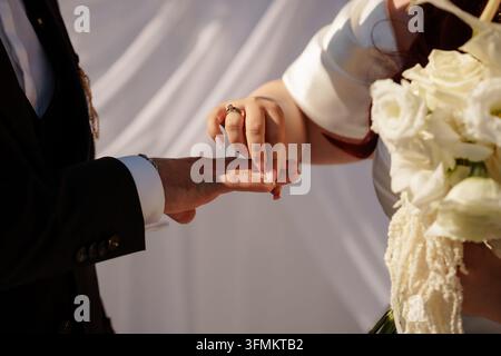 Braut, die dem Bräutigam während einer Trauungszeremonie einen goldenen Ehering an den Finger steckt, gehalten mit einem Brautstrauß, Symbol für Hingabe, Eheversprechen, ewige Liebe, Hochzeitsfeier Stockfoto