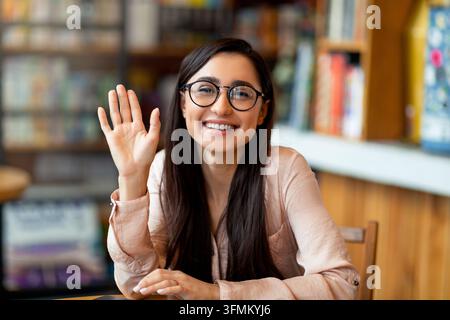 Videoanruf. Glückliche junge arabische Dame begrüßt jemanden, hat virtuelle Konferenz, winkt mit der Hand zur Kamera und lächelt, sitzt im Café. Entfernte Fernbedienung Stockfoto
