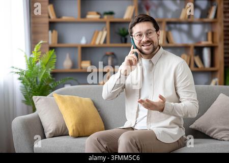 Junger bärtiger Mann in seinen 30ern lächelt während eines Telefonats, entspannt auf einem Sofa in seinem gemütlichen Wohnzimmer, lässiges Smart-Outfit, genießt einen bequemen, modernen Lebensstil Stockfoto