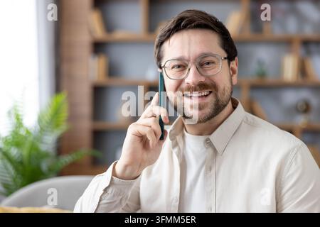 Fröhlicher bärtiger Mann mit Brille und beigem Hemd, der breit lächelt, während er ein Smartphone ans Ohr hält, und ein freundliches Telefonat zu Hause genießt, entspannt und selbstbewusst Stockfoto