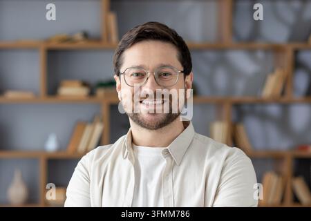 Fröhlicher männlicher Unternehmer mit Brille und Bart, der in die Kamera schaut, selbstbewusst in einem modernen Büro- oder Heimbibliotheksumfeld posiert, repräsentiert Erfolg und Professionalität Stockfoto