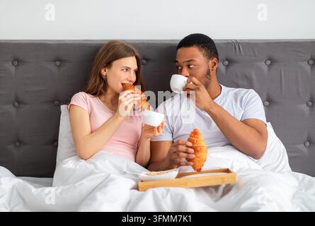 Romantisches multikulturelles Paar isst frische Croissants mit Kaffee, hat Frühstück im Bett zu Hause. Liebende junge Familie genießt eine frische, leckere Mahlzeit, verbringt Stockfoto