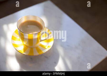 Eine gelb und weiß gestreifte Tasse, gefüllt mit Kaffee, ruht auf einem passenden Unterteller und wirft weiche Schatten, während Sonnenlicht über die weiße Marmortischplatte fällt Stockfoto
