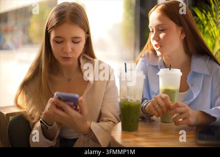 Zwei junge Frauen sitzen zusammen an einem Holztisch im Café, eine verwendet ein Smartphone, während die andere ein eisgekühltes grünes Getränk in einem hellen Innenraum hält. Matcha Eve Stockfoto