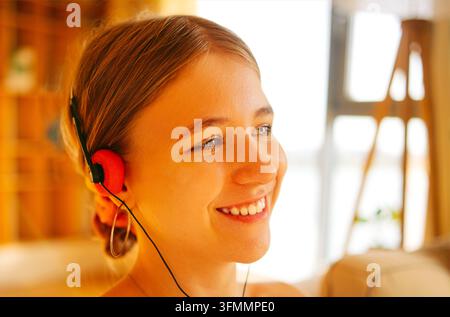 Lächelnde Frau mit braunem Haar trägt rote und schwarze Kopfhörer, während warmes Sonnenlicht über ihr Gesicht fällt, neben hellen Fenstern und Möbeln. Analog Revi Stockfoto