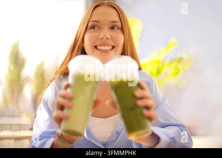 Lächelnde rothaarige Frau präsentiert zwei grüne Smoothies in Richtung Kamera, mit hellem Blumendekor und einem sanft unscharfen, sonnenbeschienenen Innenraum im Hintergrund. Mat Stockfoto