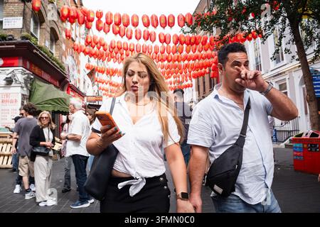 FRAU NUTZT HANDY WÄHREND DES GEHENS MIT PARTNER, LONDON CHINATOWN, REISEBERICHT, LONDON, ENGLAND: Eine blondhaarige Frau ist in ihr Handy vertieft, während sie mit einem Mann auf der Macclesfield Street in Londons Chinatown unter festlichen roten Laternen spaziert. INFO: Londons Chinatown, eingebettet im pulsierenden Herzen von Soho, ist ein geschäftiger kultureller Mittelpunkt, berühmt für seine authentischen asiatischen Restaurants, traditionellen Bäckereien und neonbeleuchteten Straßen. Die ikonischen roten Laternen bieten eine perfekte Kulisse für Feinschmecker. Stockfoto