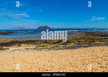 Pittoreske Küstenansicht von sandigem Strand und felsigem vulkanischem Riff bei Ebbe mit den Inseln Lobos und Lanzarote am Horizont in Corralejo, Fuerteventura, Ca Stockfoto