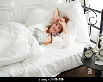 Junge Frau mit langen roten Haaren in einem weißen Pyjama liegt im Bett unter einer Decke und lächelt. Morgen. Stockfoto