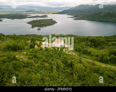 Luftaufnahme des Slansko-Sees mit einem kleinen Steinhaus auf einem üppigen grünen Hügel, umgeben von dichtem Wald und Bergen in der Gemeinde Niksic, Montenegro Stockfoto