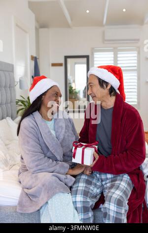 Senior asiatischer Mann und afrikanische Frau, die ein eingewickeltes Geschenk austauschen, sitzen auf dem Bett in Weihnachtsmützen Weihnachten Stockfoto