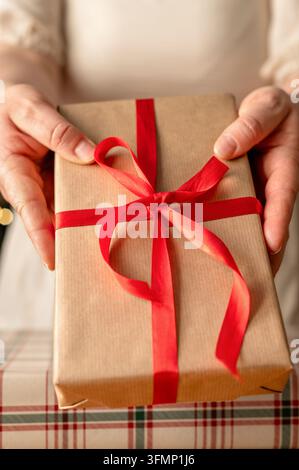 Nahaufnahme von Händen, die ein in braunes Kraftpapier eingewickeltes Weihnachtsgeschenk mit einer leuchtend roten Schleife über einer festlichen karierten Tischdecke halten. Stockfoto