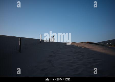 Fußabdrücke kreuzen schattige Sanddünen neben verwitterten Zaunpfählen an der Atlantikküste Portugals unter einem klaren blauen Himmel und warmer Tiefluft Stockfoto