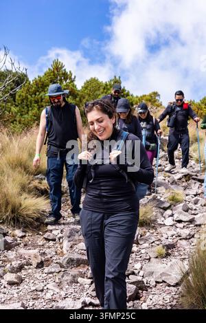 Lächelnde Frau führt eine Gruppe von Wanderern, die einen felsigen Bergpfad hinabsteigen, mit Rucksäcken und Trekkingstöcken durch trockenes Gras und Kiefern unterhalb Stockfoto