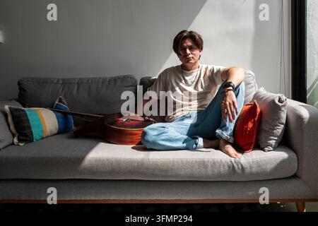 Junger männlicher Musikkomponist sitzt barfuß auf einem grauen Sofa neben einer Akustikgitarre, ruht im Sonnenlicht in einem modernen Zuhause mit dramatischen Schatten. Stockfoto