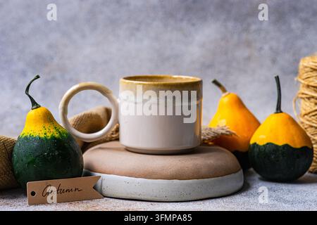 Gemütliche Herbsttischdekoration mit einer handgefertigten Keramiktasse, drei dekorativen Kürbissen, Leinenstoff und einem handgeschriebenen saisonalen Schild auf einem neutralen Textil Stockfoto