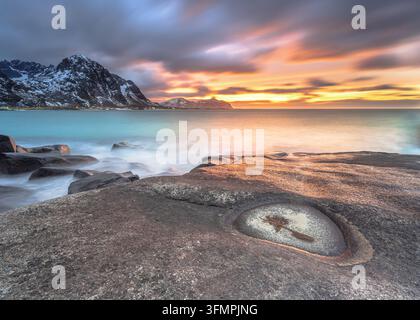 Felsenküste, die im letzten Sonnenlicht in einem arktischen Fjord mit vorbeiziehenden Wolken am Vareid Strand Beach, Flakstad leuchtet. Atemberaubender Sonnenuntergang mit Bergen am Horizont o Stockfoto