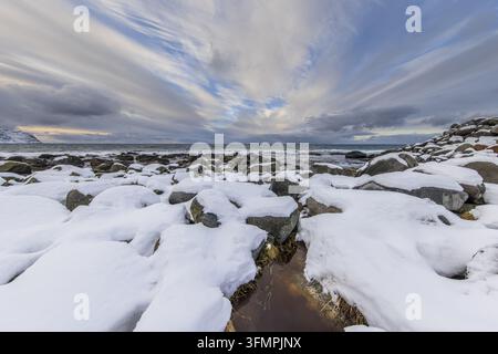 Felsenküste mit im letzten Sonnenlicht schneebedeckten Gipfeln in einem arktischen Fjord mit vorbeiziehenden Wolken am Vikten Strand Beach, Vareid, Flakstad. Atemberaubender Sonnenuntergang mit Bergen Stockfoto