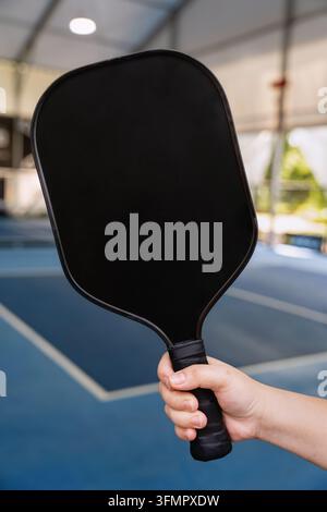 Hand, die einen leeren schwarzen Pickleball-Schläger hält, bereit zum Spiel auf einem Platz. Stockfoto
