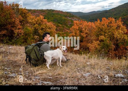 Ein Mann sitzt auf trockenem Gras mit einem Hund neben sich. Die Landschaft hat farbenfrohe Bäume und Hügel im Hintergrund. Stockfoto