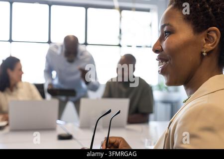 Afroamerikanische Frau hält eine Brille, lächelt, schaut auf das Team im Büro mit Laptops, Kopierraum Stockfoto