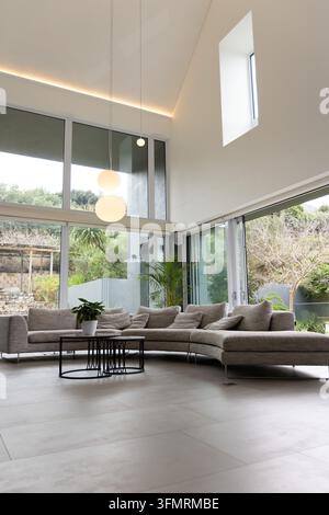 Gekrümmtes graues Ecksofa zentriert in einem modernen Zuhause, zeigt Kissen und schwarzen Couchtisch. Minimalistisch, zeitgenössisch, sonnenbeschienen, Verglasung, Grünpflanzen, Pendelleuchte, Sofa Stockfoto
