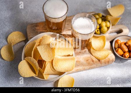 Zwei Gläser schaumiges Bier mit Kartoffelchips, grünen Oliven und kleinen Würstchen, die auf einem rustikalen Holzbrett arrangiert sind und eine lässige Vorspeisenauswahl bilden Stockfoto