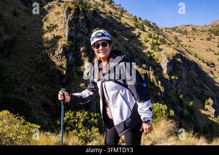 Lächelnde Frau mit Helm und Sonnenbrille macht eine Pause auf einer Bergwanderung, hält einen Trekkingstock neben schroffen Klippen, Buschvegetation und einem sonnenbeschienenen Tal Stockfoto