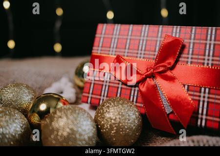 Nahaufnahme eines Feiertagsstilleben mit einer rot karierten Geschenkschachtel, Satin-Schleife und schimmernden goldenen Weihnachtskugeln, ideal für festliche Werbung, saisonal Stockfoto