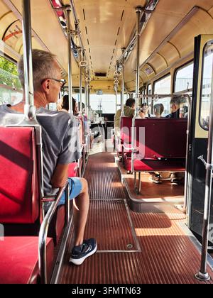 Innenansicht von Passagieren, die in einem Vintage-ÖPNV-Bus fahren, zeigt rote Retro-Sitzplätze, den Mittelgang und Handläufe an einem sonnigen Tag. Stockfoto