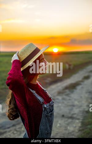 Junge Frau mit Strohhut bei Sonnenuntergang. Junge Frau trägt einen Strohhut und steht auf einer ländlichen Straße während eines warmen Sonnenuntergangs auf dem Land. Stockfoto