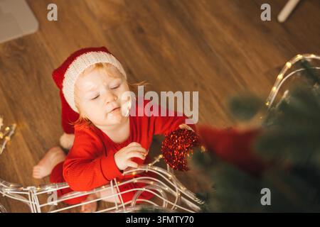 Niedliches Kleinkind mit Weihnachtsmannmütze spielt mit Weihnachtsdekoration Stockfoto